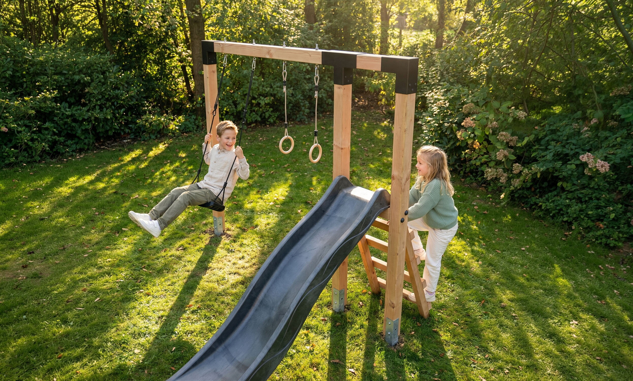 Zwei Kinder spielen im Garten auf einem Holzspielger&auml;t mit Schaukel, Ringen und schwarzer Rutsche.